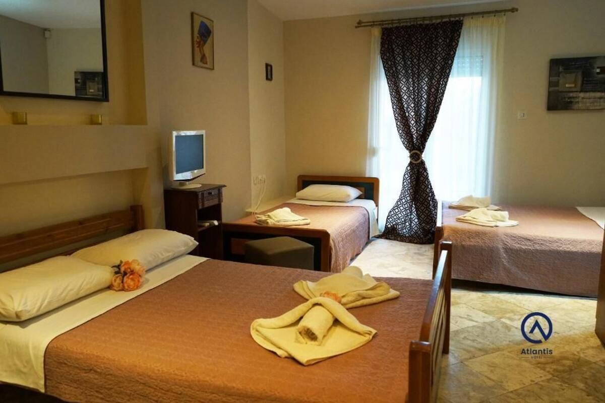 Ett hotellrum med tre sängar, en tv, ett skrivbord och ett fönster med mönstrade gardiner.