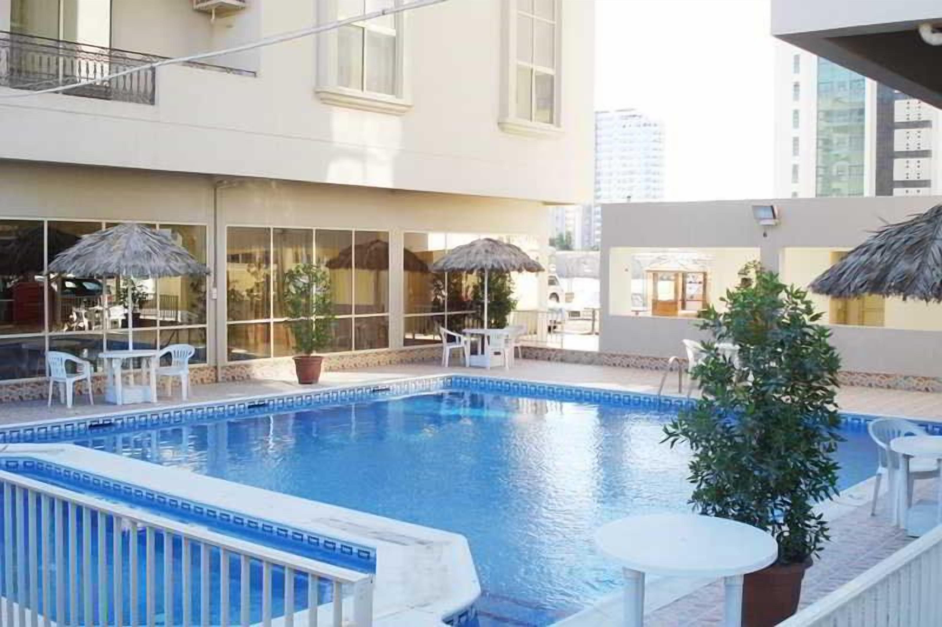An indoor swimming pool with a tiled edge, surrounded by lounge chairs and umbrellas, with a building in the background.