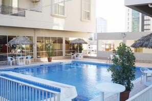 An indoor swimming pool with a tiled edge, surrounded by lounge chairs and umbrellas, with a building in the background.