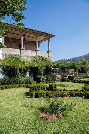Un edificio con un porche cubierto, columnas de piedra y un jardín con setos y flores.