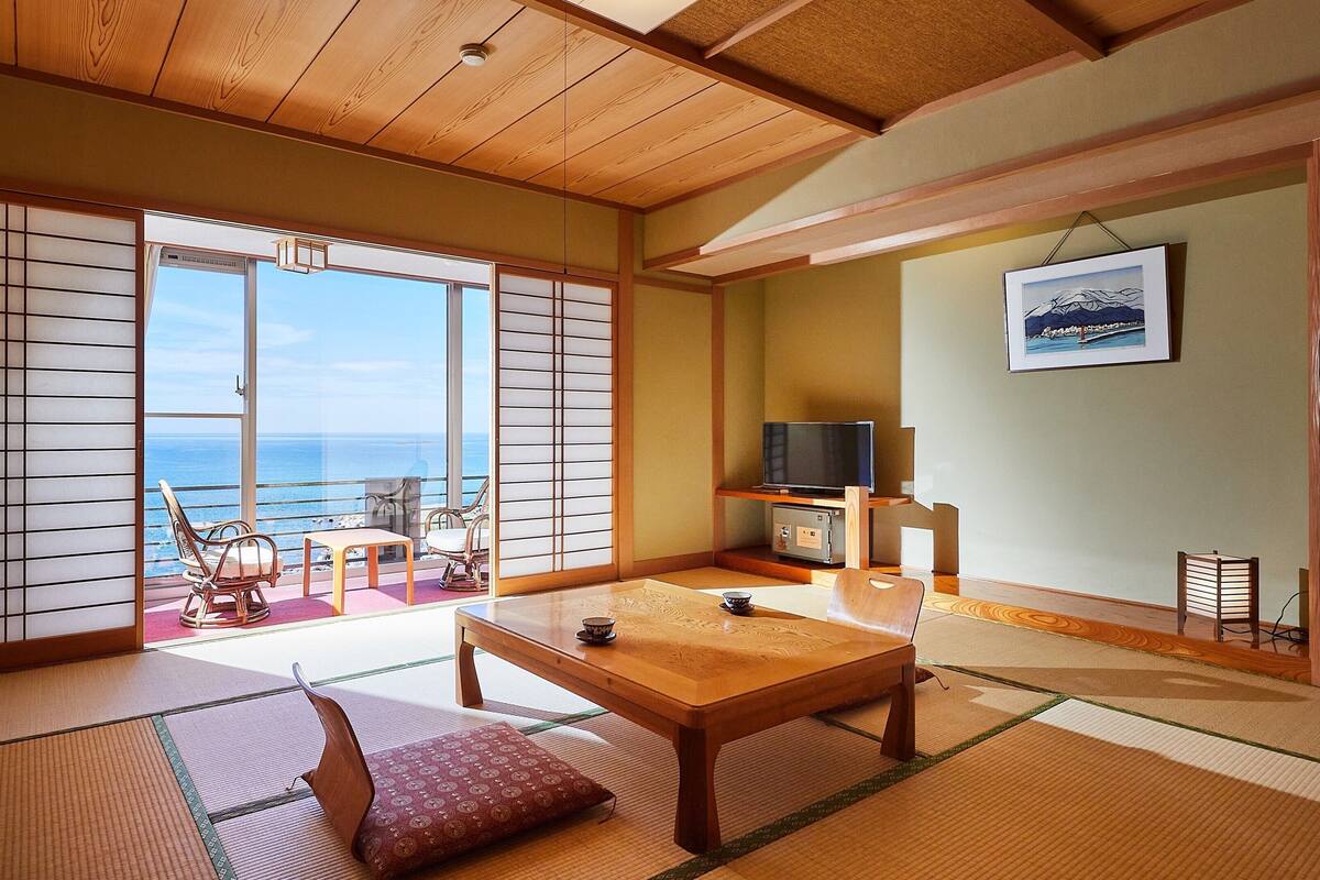 Una habitación tradicional japonesa con suelo de tatami, una puerta corredera con vistas al mar, un televisor y un cuadro en la pared.