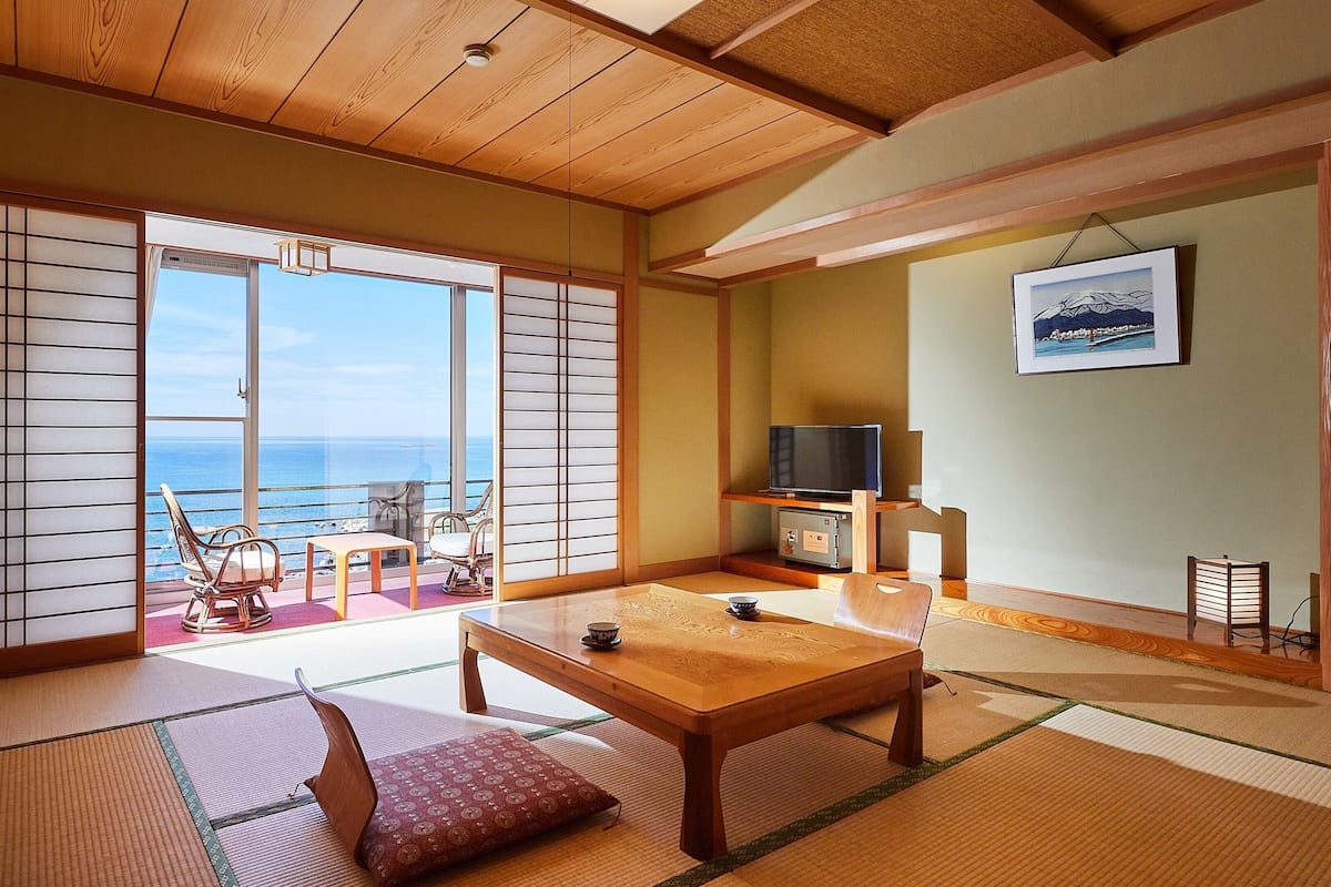 A traditional Japanese room with tatami flooring, a sliding door with a view of the sea, a television, and a framed picture on the wall.