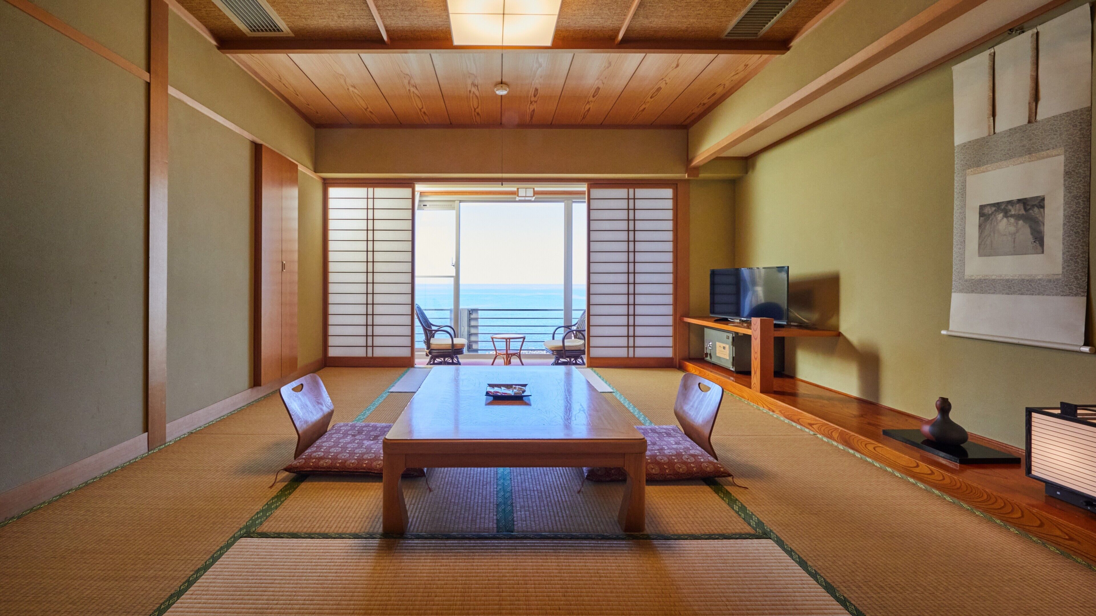 A traditional Japanese room with tatami flooring, a low table, and sliding doors leading to a balcony with a view of the sea.