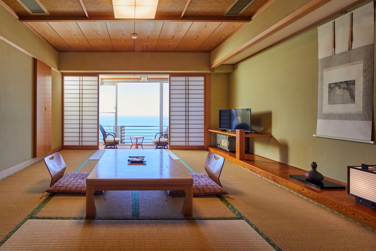 Una habitación japonesa tradicional con suelo de tatami, una mesa baja y puertas correderas que dan a un balcón con vistas al mar.