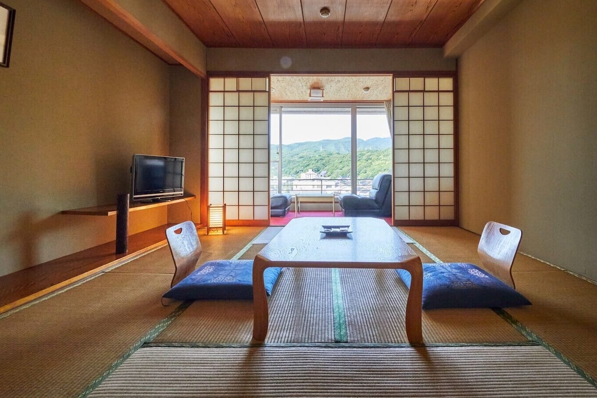 Una habitación tradicional japonesa con suelo de tatami, una mesa baja, cojines, un televisor y vistas a la montaña.