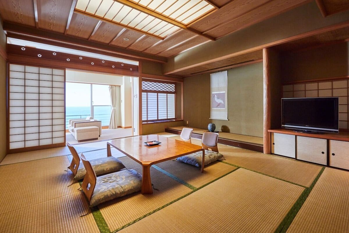 Una habitación tradicional japonesa con suelo de tatami, una mesa baja, sillas, un televisor y puertas correderas.