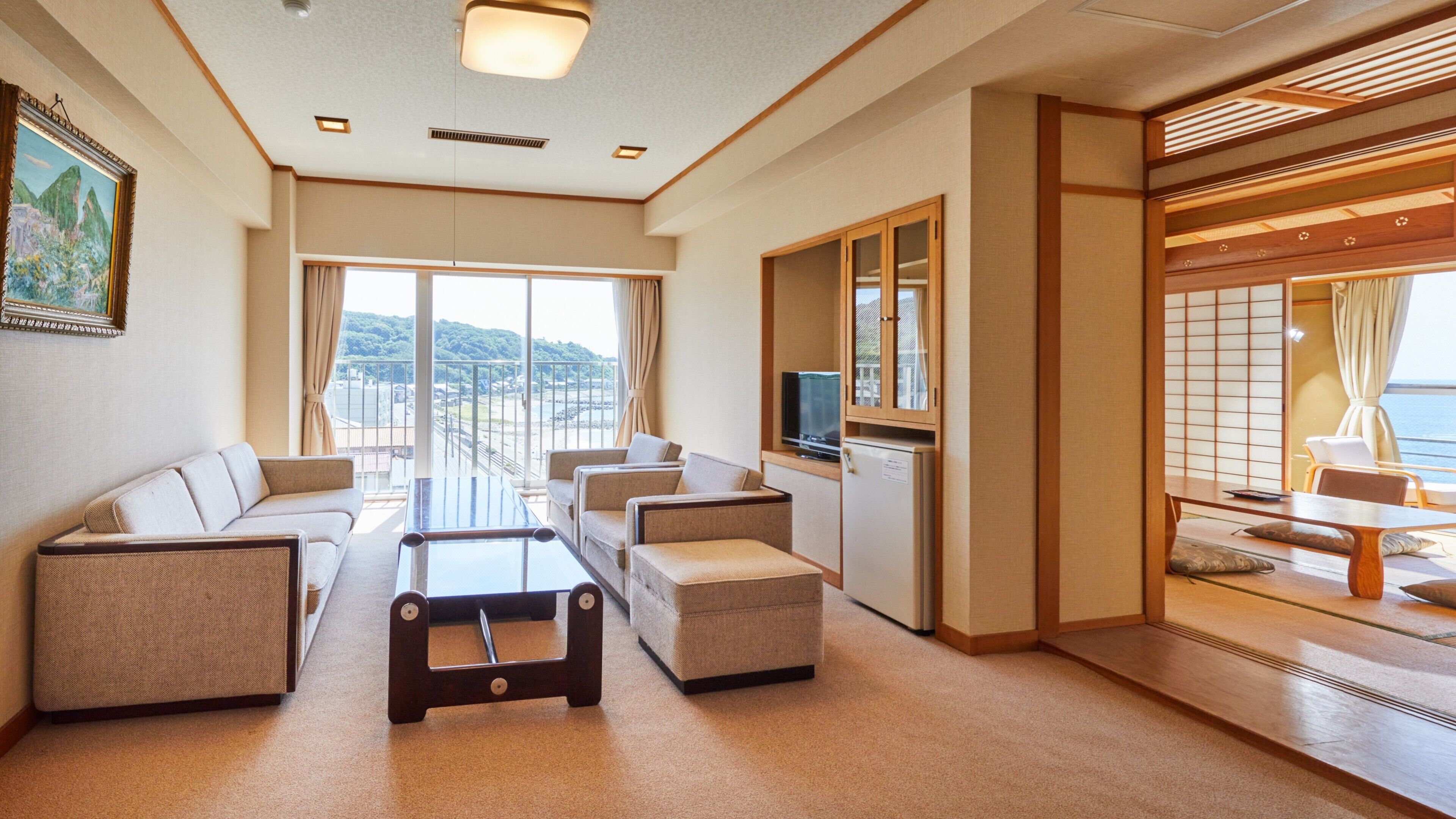 A hotel room with a sofa, armchairs, a coffee table, a TV, and a sliding door leading to a balcony with a view.