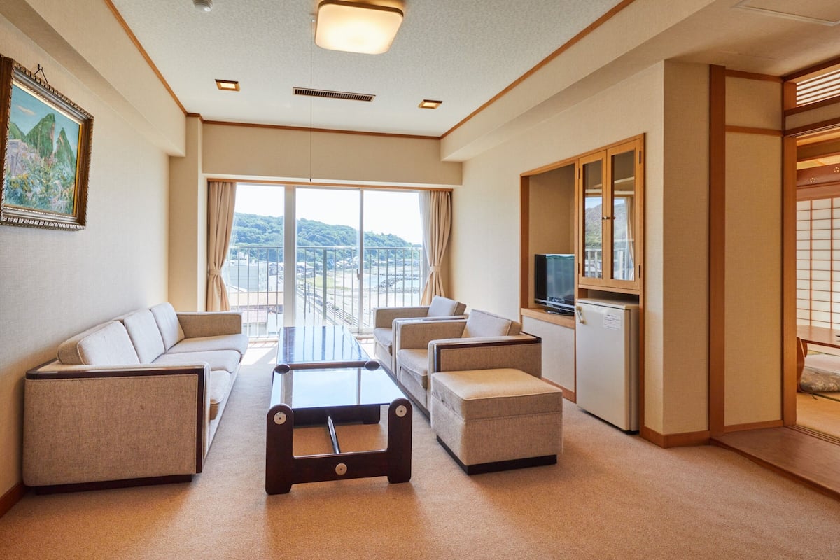 Habitación de hotel con sofá, sillones, una mesa de centro, televisión y puerta corrediza que da a un balcón con vistas.