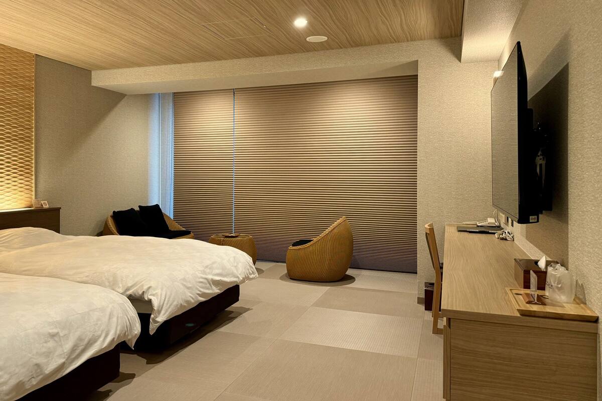 Una habitación de hotel moderna con dos camas, un televisor de pantalla plana, techo de madera y un ventanal grande con persianas.