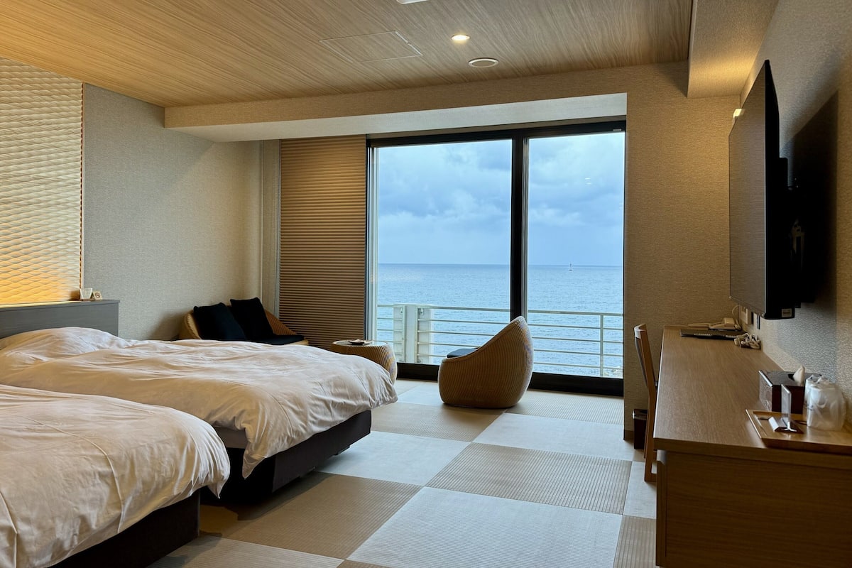 Habitación de hotel con dos camas, un ventanal con vistas al mar, un televisor de pantalla plana y piso de madera.