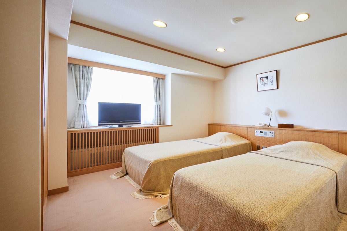 Habitación de hotel con dos camas, televisión, escritorio con lámpara y ventana con cortinas.