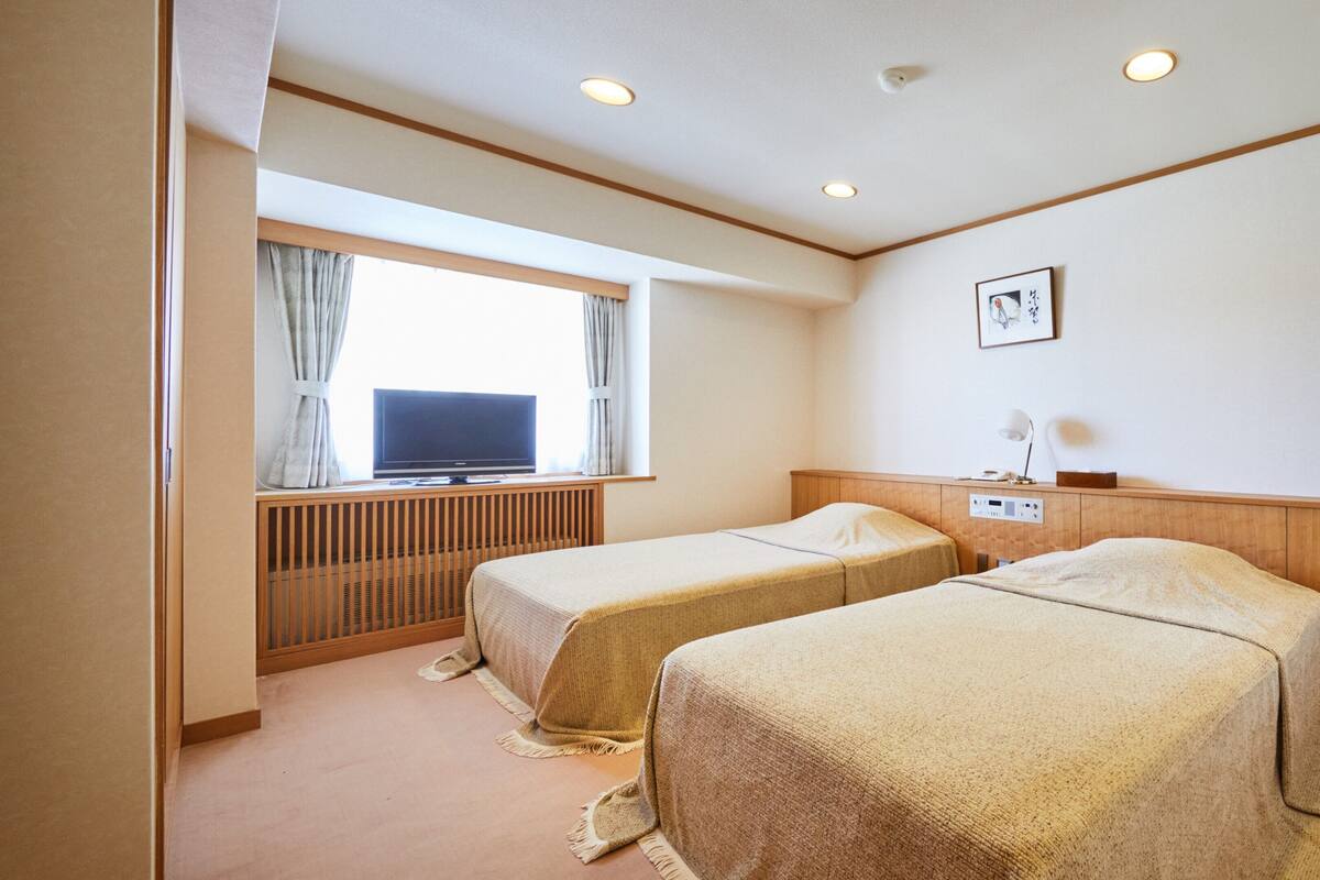 A hotel room with two beds, a TV, a desk with a lamp, and a window with curtains.