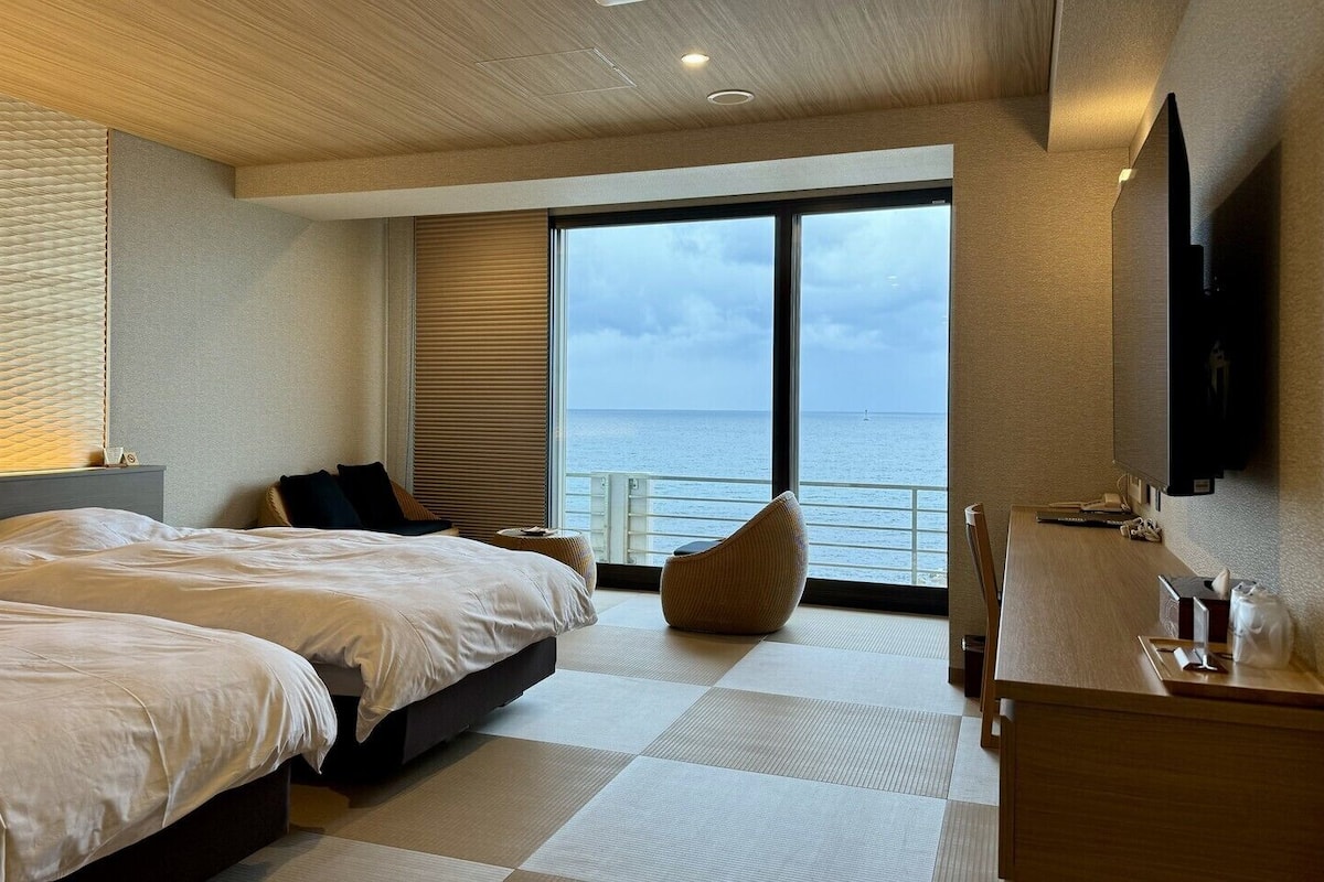 Una habitación de hotel moderna con dos camas, un ventanal con vistas al mar, un televisor de pantalla plana y un escritorio de madera.