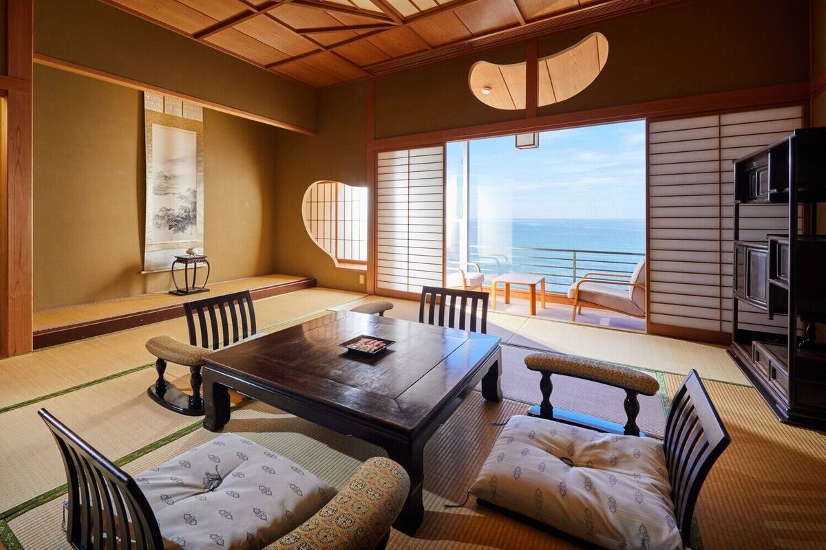 Una habitación japonesa tradicional con suelo de tatami, una mesa baja y sillas, un ventanal con vistas al mar y un puertas correderas.