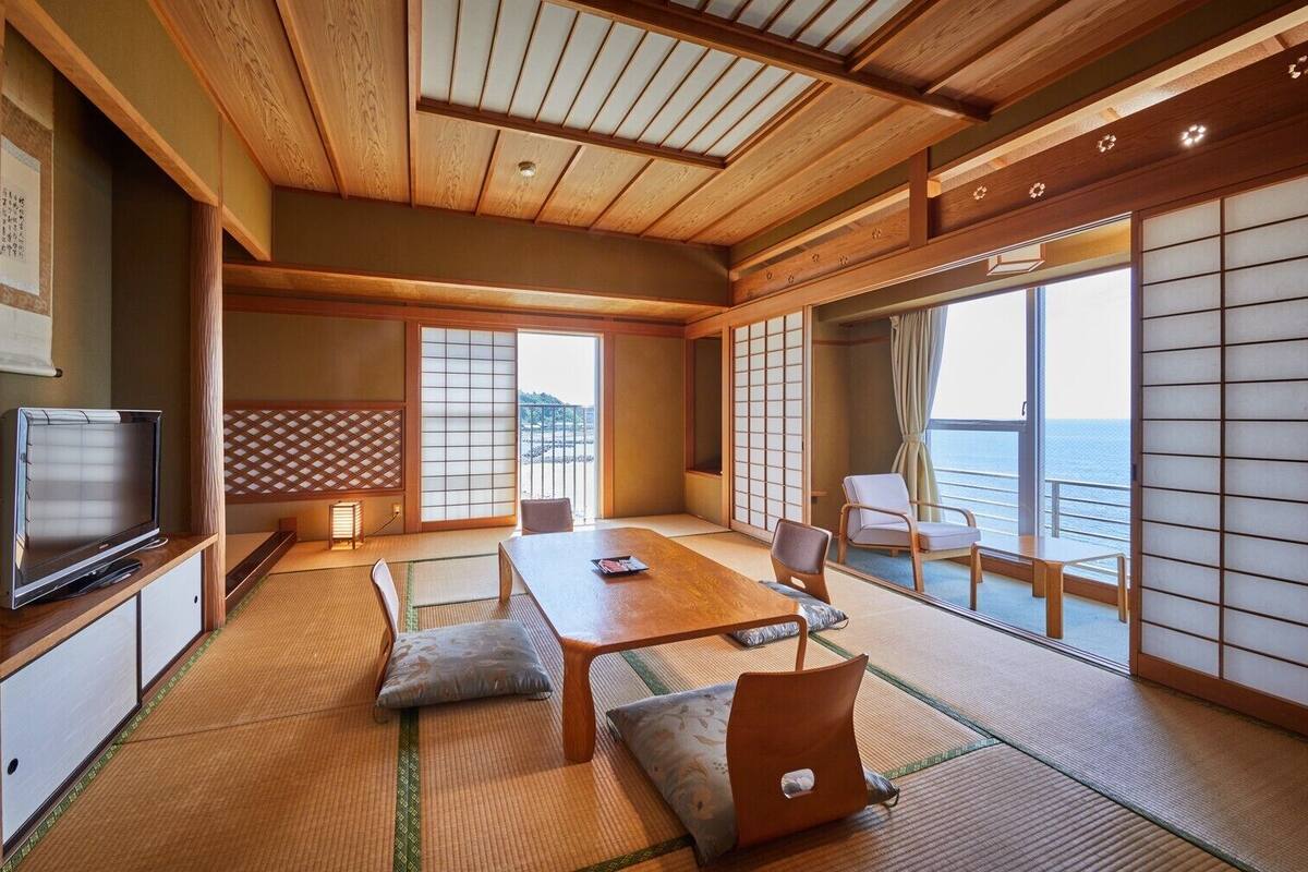Una habitación tradicional japonesa con suelo de tatami, una mesa baja, cojines, un televisor y puertas correderas con vistas al mar.