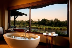 A bathroom with a large bathtub, a window with a view of a river and greenery, and a small table with toiletries. - Lion Sands Narina Lodge (Mpumalanga-SKUKUZA)