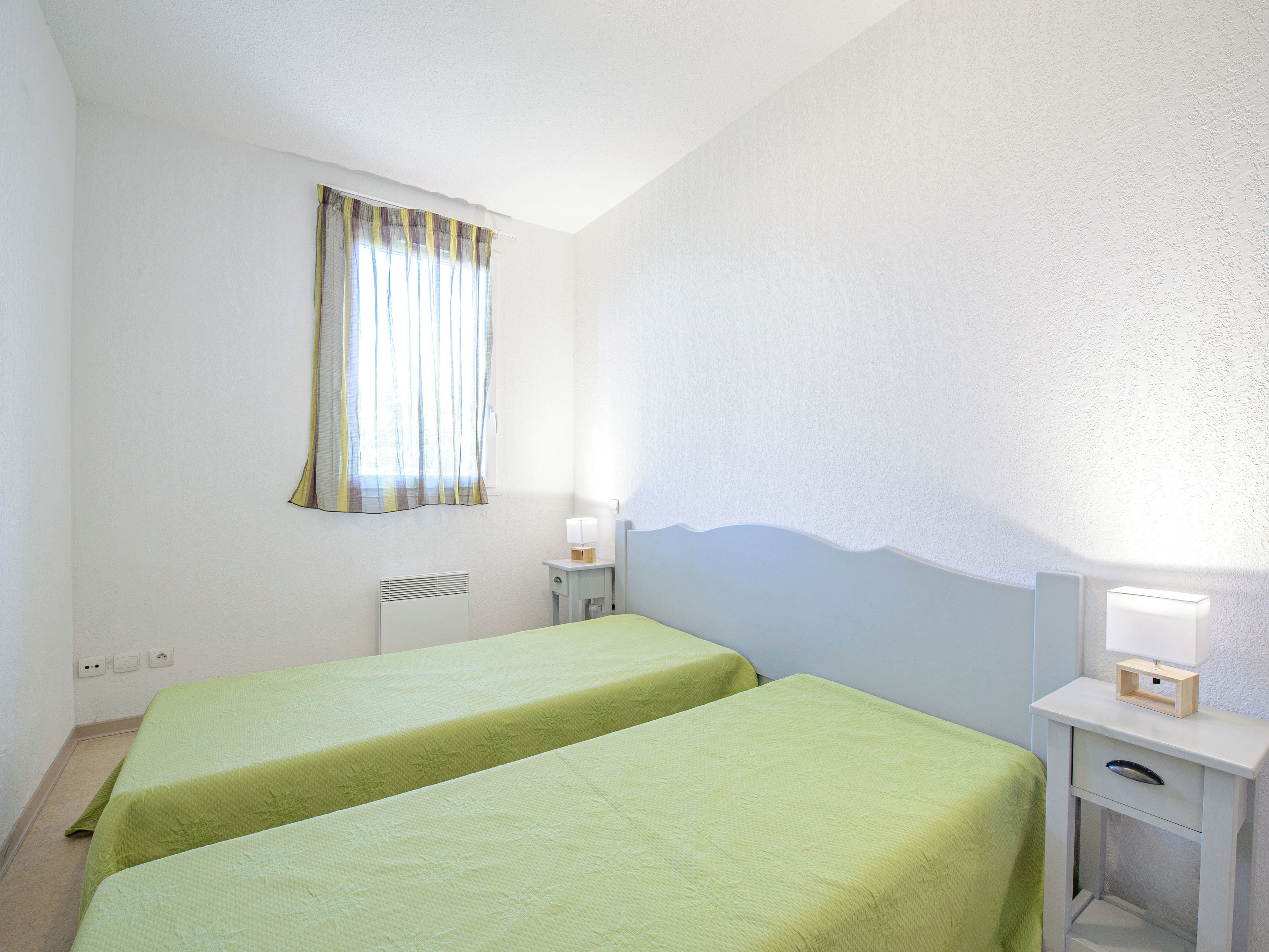 Una habitación con dos camas, una ventana con cortinas, una mesita de noche con lámpara y un radiador.