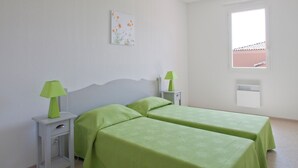 A room with two beds, each with a green bedspread, two bedside tables with lamps, a white wall, a window, and a whiteboard.