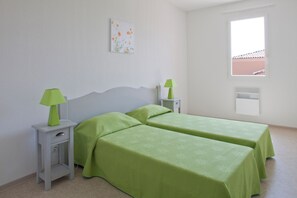 Una habitación con dos camas, cada una con colcha verde, dos mesitas de noche con lámparas, una pared blanca, una ventana y una pizarra blanca.