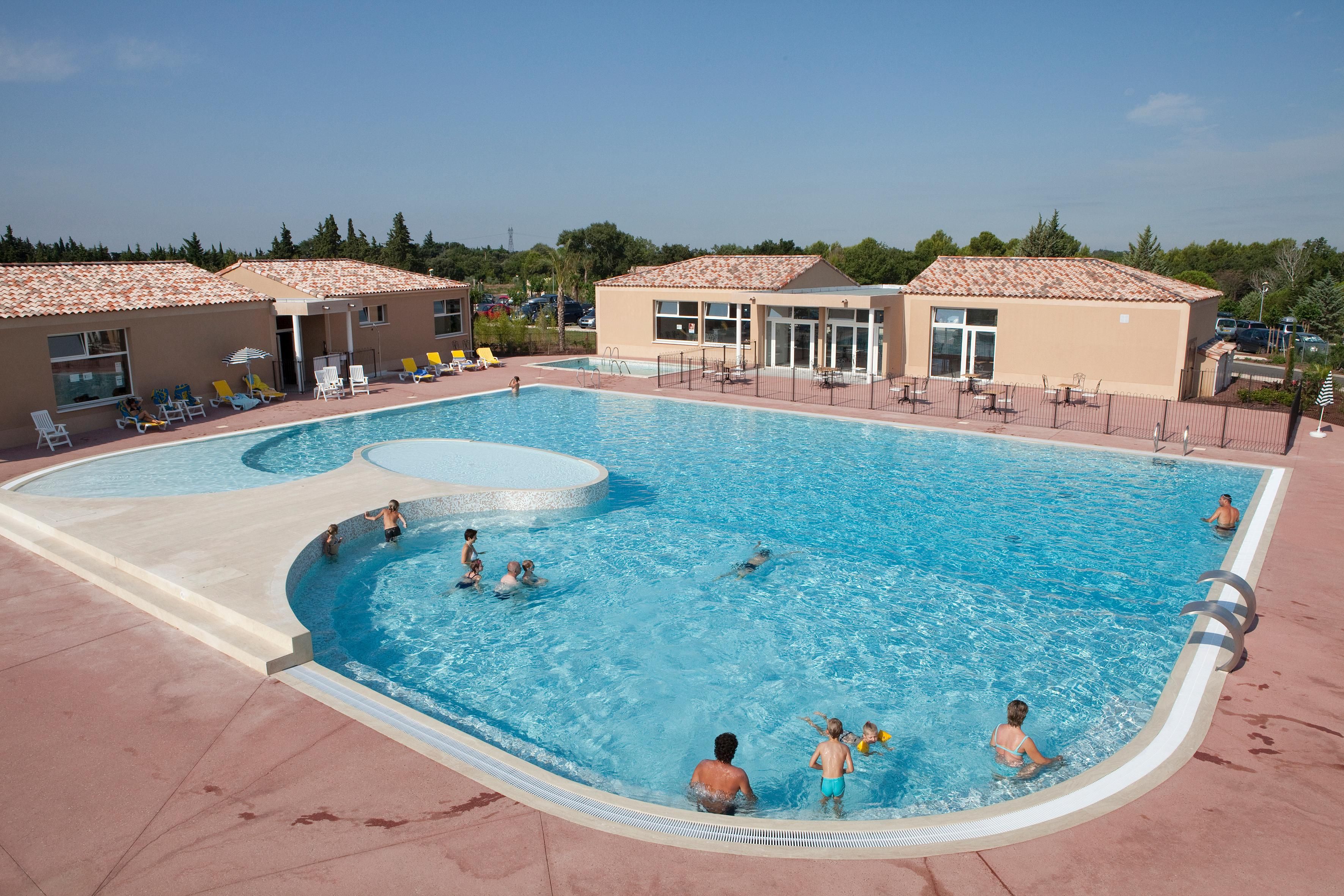 Una piscina con múltiples carriles, rodeada de tumbonas y sillones de descanso, con edificios al fondo.