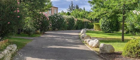 Un chemin de gravier bordé d’une végétation luxuriante et de gros rochers, qui mène à un bâtiment à la façade de couleur claire.
