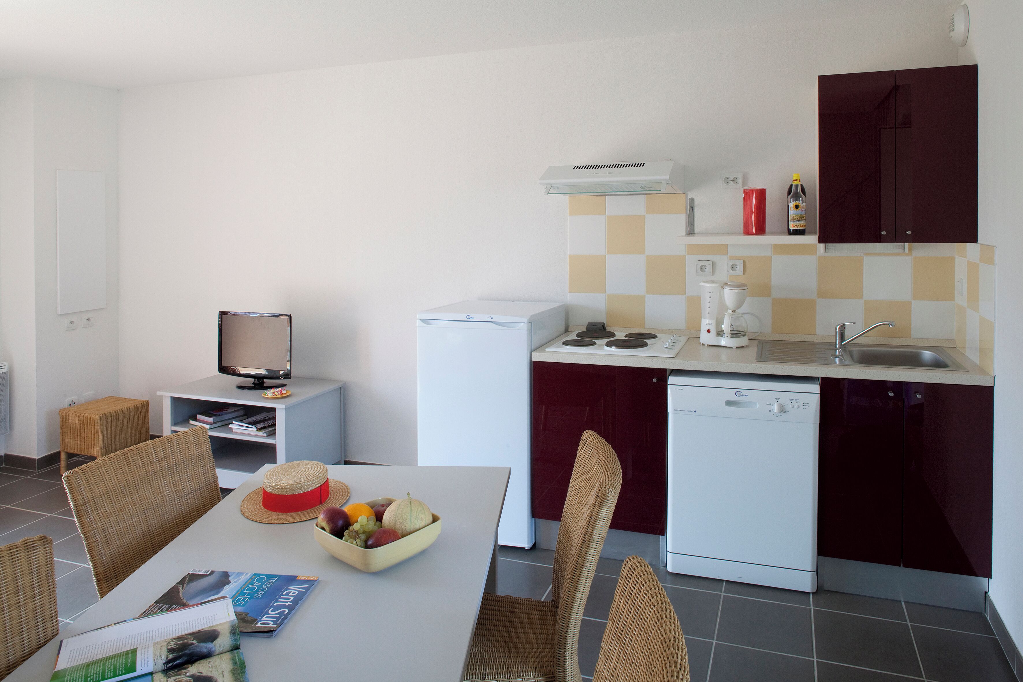 Una cocina compacta con lavavajillas, cafetera y microondas. Hay una mesa de comedor con un tazón de fruta y una revista.