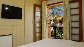 Un dormitorio con un televisor montado en la pared, una cama con cubrecama blanco y vistas a un espacio exterior con un árbol y un edificio, visible a través de una puerta de madera con cortina.