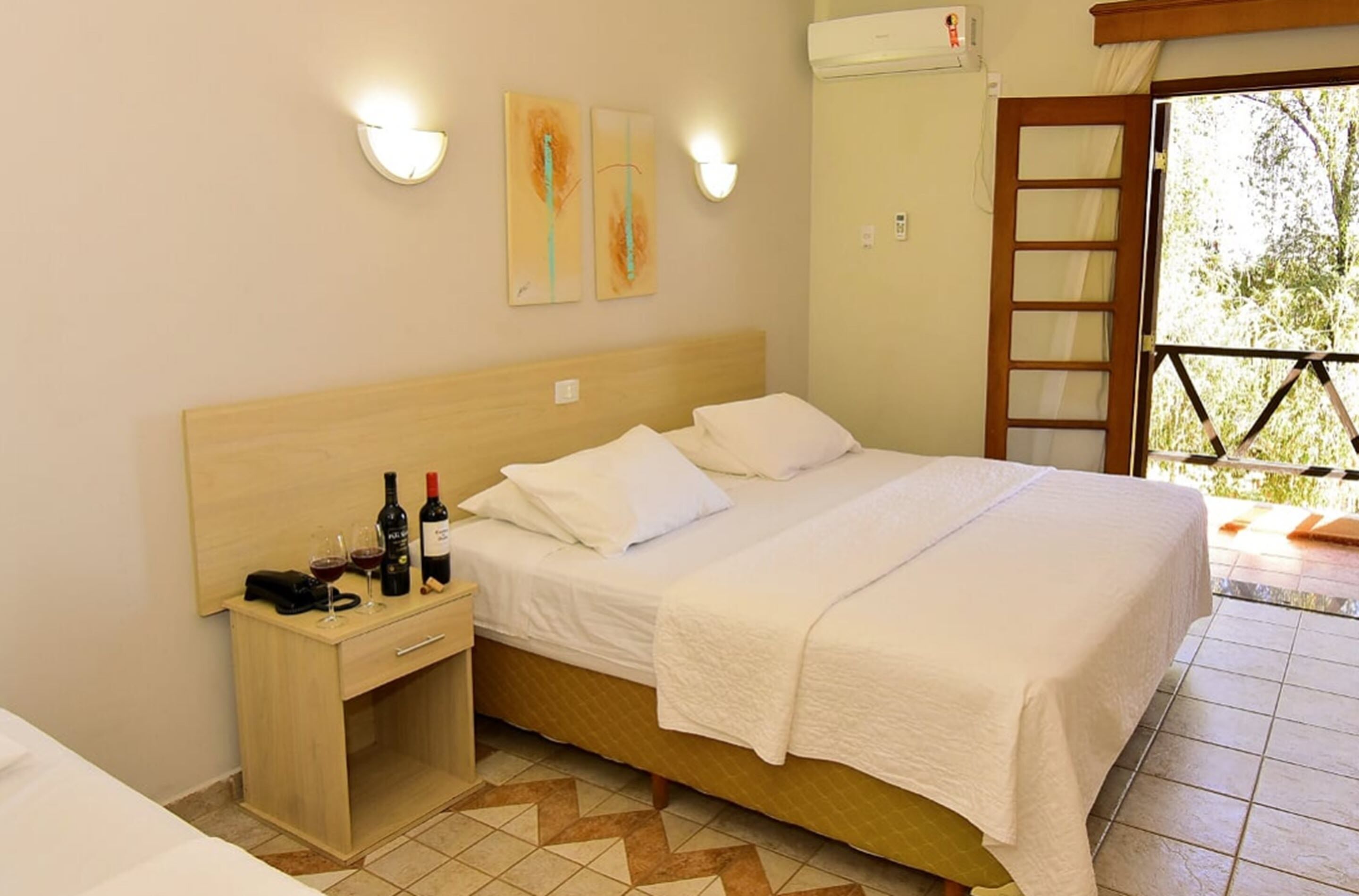 A hotel room with a wooden bed, white linens, a bedside table with wine bottles, and a view of greenery through a door.