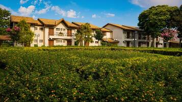 Uma fileira de edifícios modernos, com vários andares e varandas, rodeada por um jardim bem cuidado com flores amarelas.