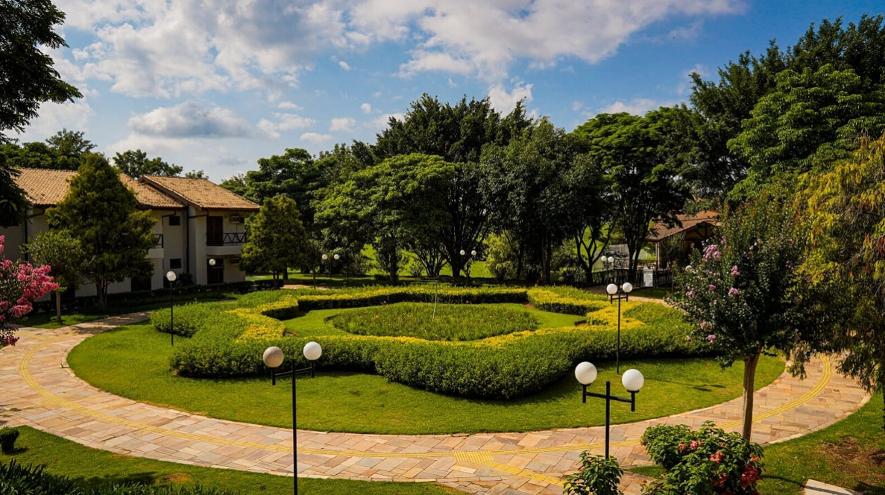 A landscaped garden with a circular pathway, neatly trimmed hedges, and a building with a tiled roof in the background.