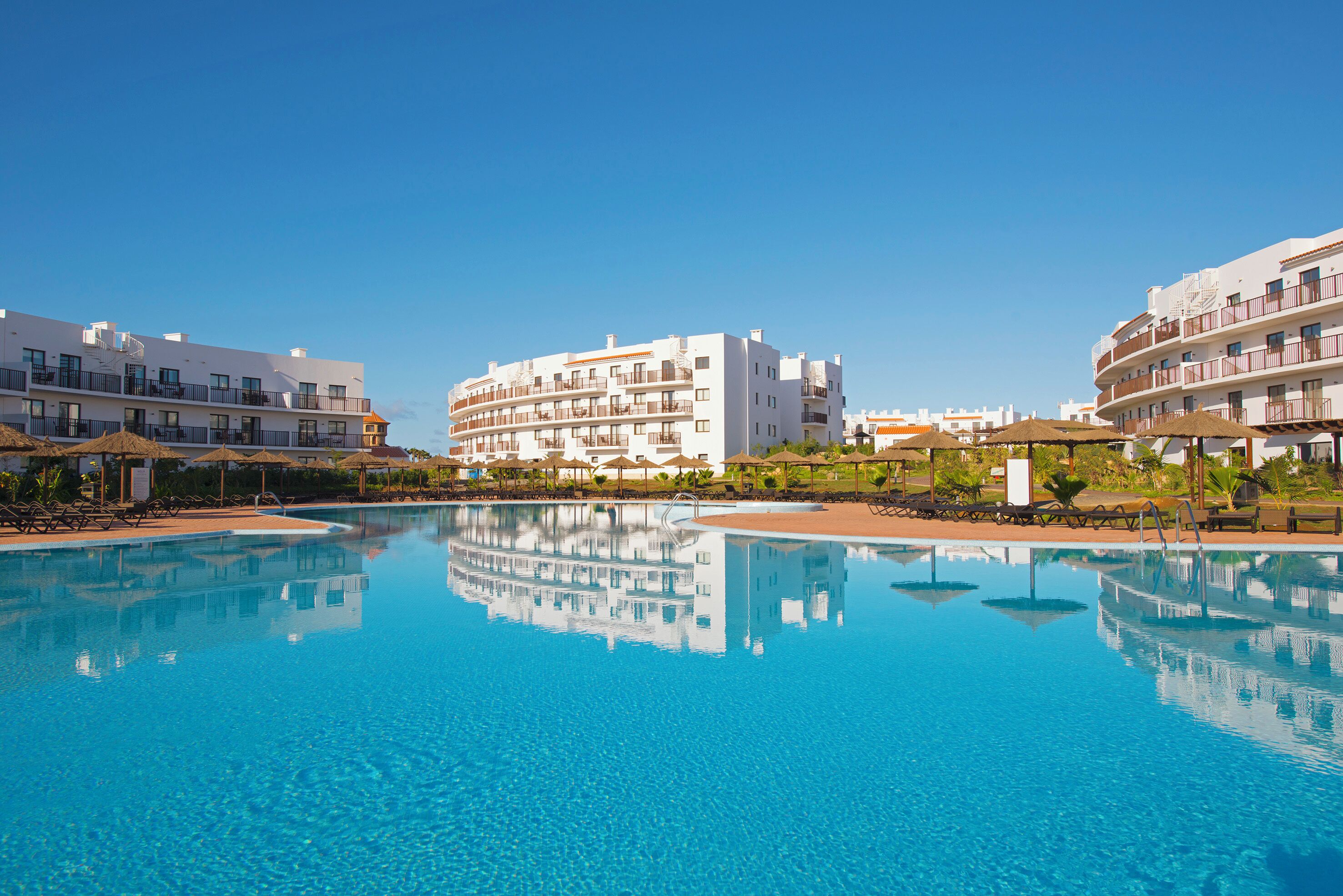 A swimming pool with clear blue water, surrounded by white buildings with balconies and sun loungers.
