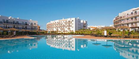 A swimming pool with clear blue water, surrounded by white buildings with balconies and sun loungers.