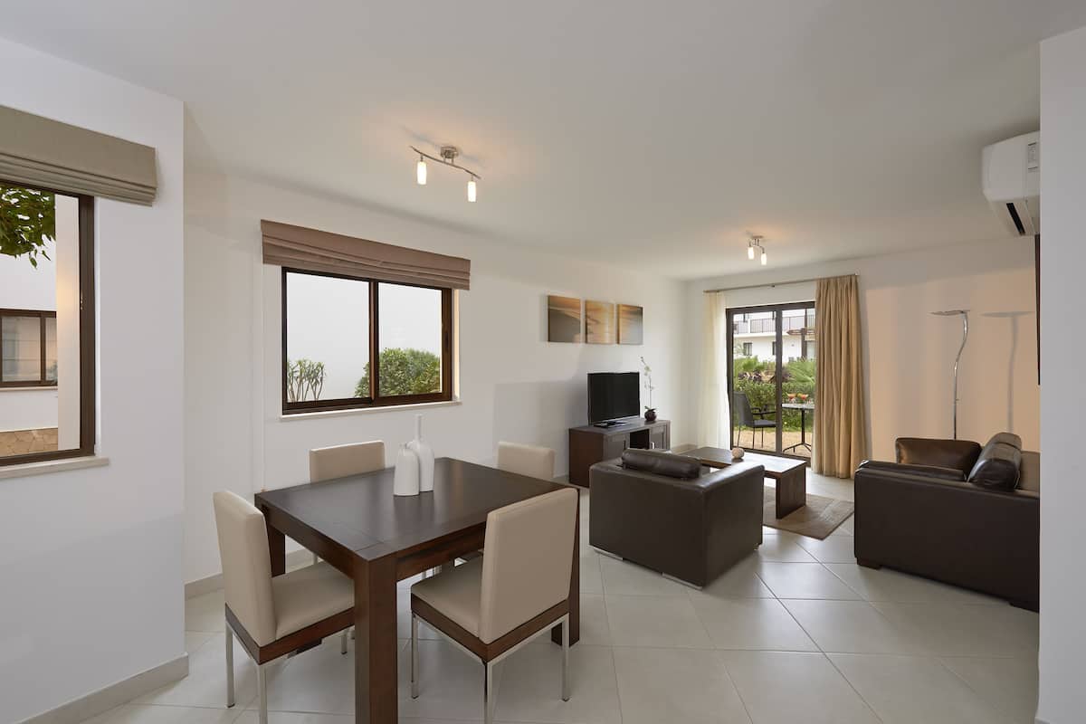 A modern living room with a dining area, a flat-screen TV, and a balcony with curtains.