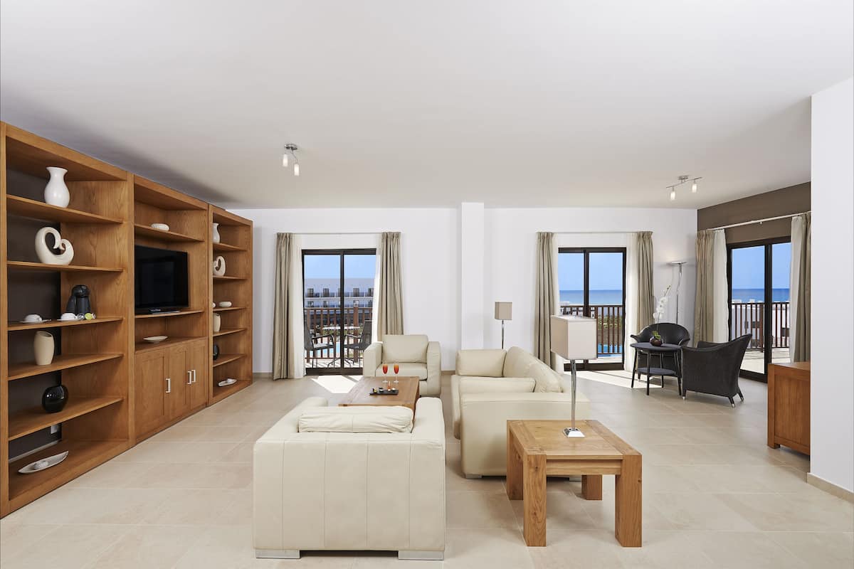 A spacious living room with a sofa, coffee table, and wooden shelving unit. There are large windows with curtains, a balcony with outdoor seating, and a television on a wooden stand.