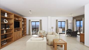 Amplia sala de estar con sofá, mesa de centro y estantería de madera. Cuenta con ventanas amplias con cortinas, un balcón con mobiliario exterior y un televisor sobre un mueble de madera.