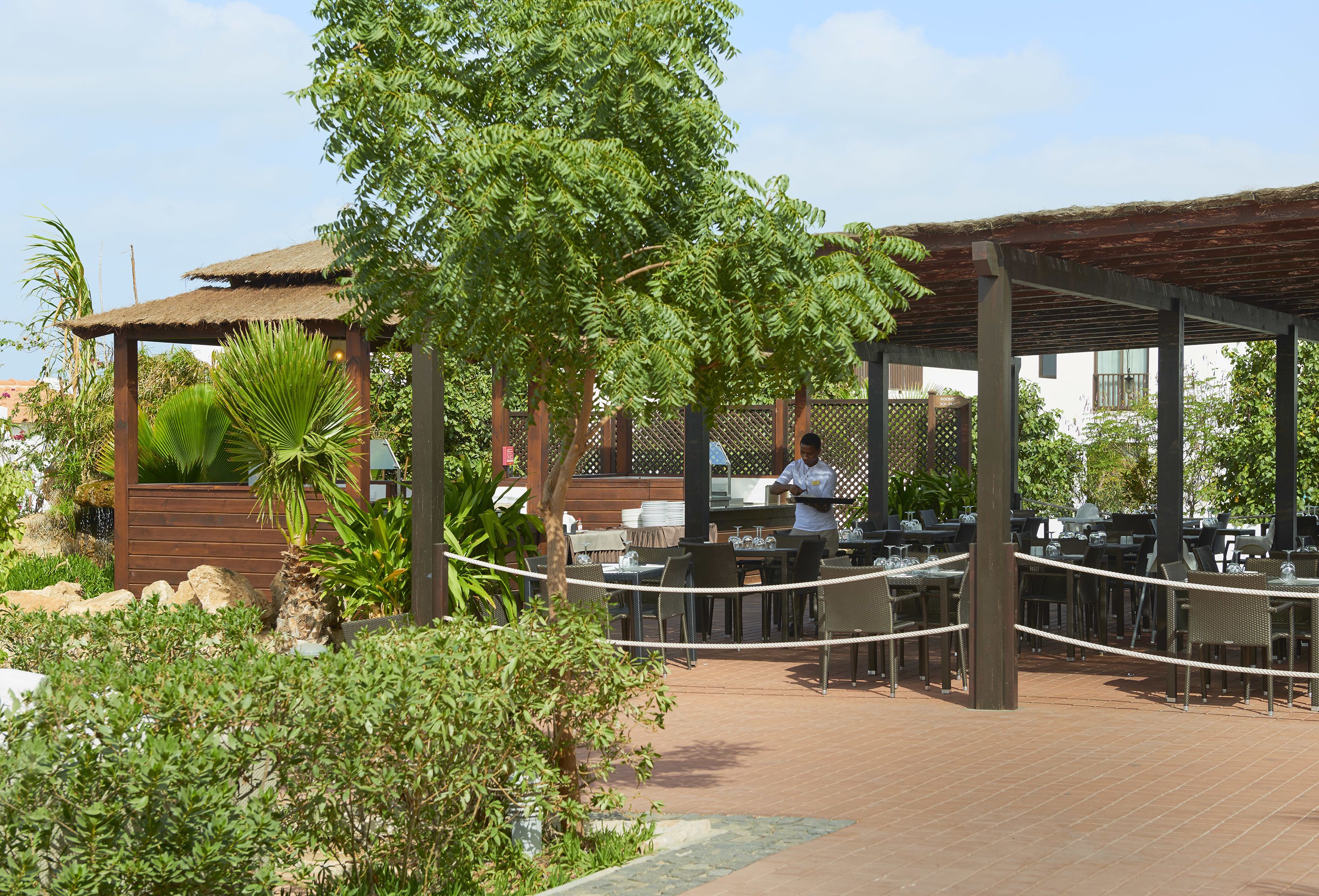 Un espacio de comedor al aire libre con pérgolas de madera, mesas y sillas, rodeado de follaje y una estructura de techo de paja.