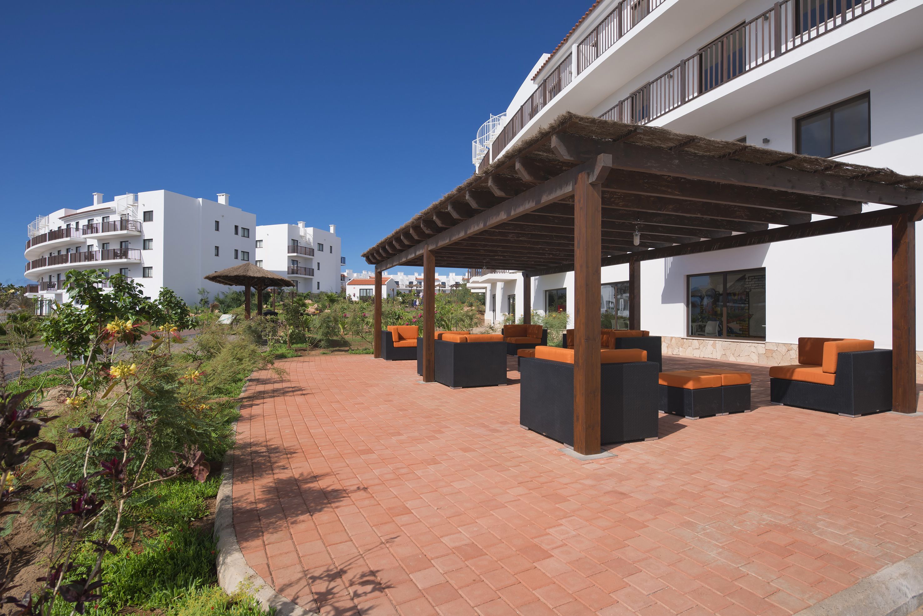 A covered outdoor seating area with wooden pillars and orange cushions, surrounded by a brick patio and greenery.