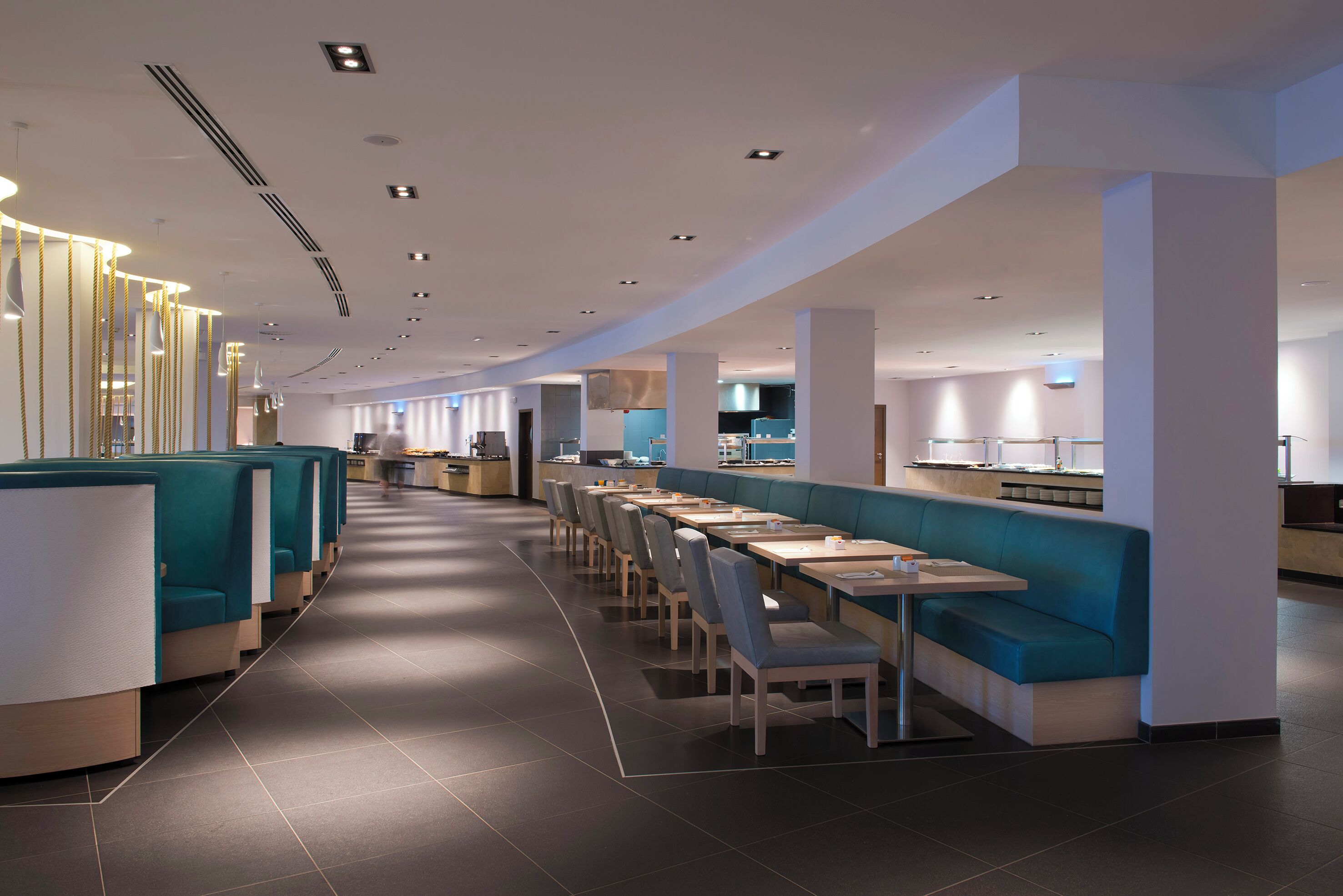 A modern restaurant with long communal tables and booths, a clean tiled floor, and recessed lighting.