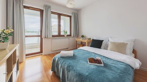 A bedroom with a bed, a nightstand, a window with curtains, a radiator, and a view of buildings outside.