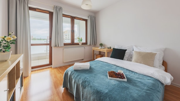 Una camera da letto con un letto, un comodino, una finestra con tende, un termosifone e vista sugli edifici.