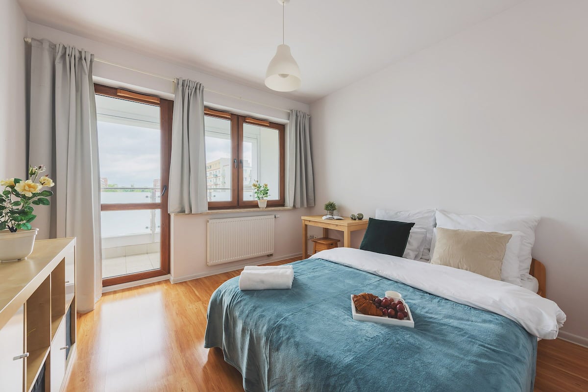A bedroom with a bed, a bedside table, a window with curtains, and a view of buildings.