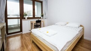 A bedroom with a wooden bed, a desk, and a chair. There is a window with curtains and a view of buildings outside.