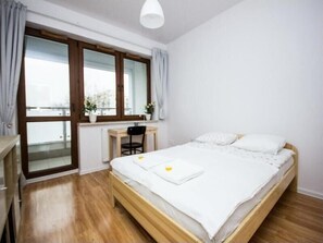 Un dormitorio con una cama de madera, un escritorio y una silla. Hay una ventana con cortinas y vistas a edificios.