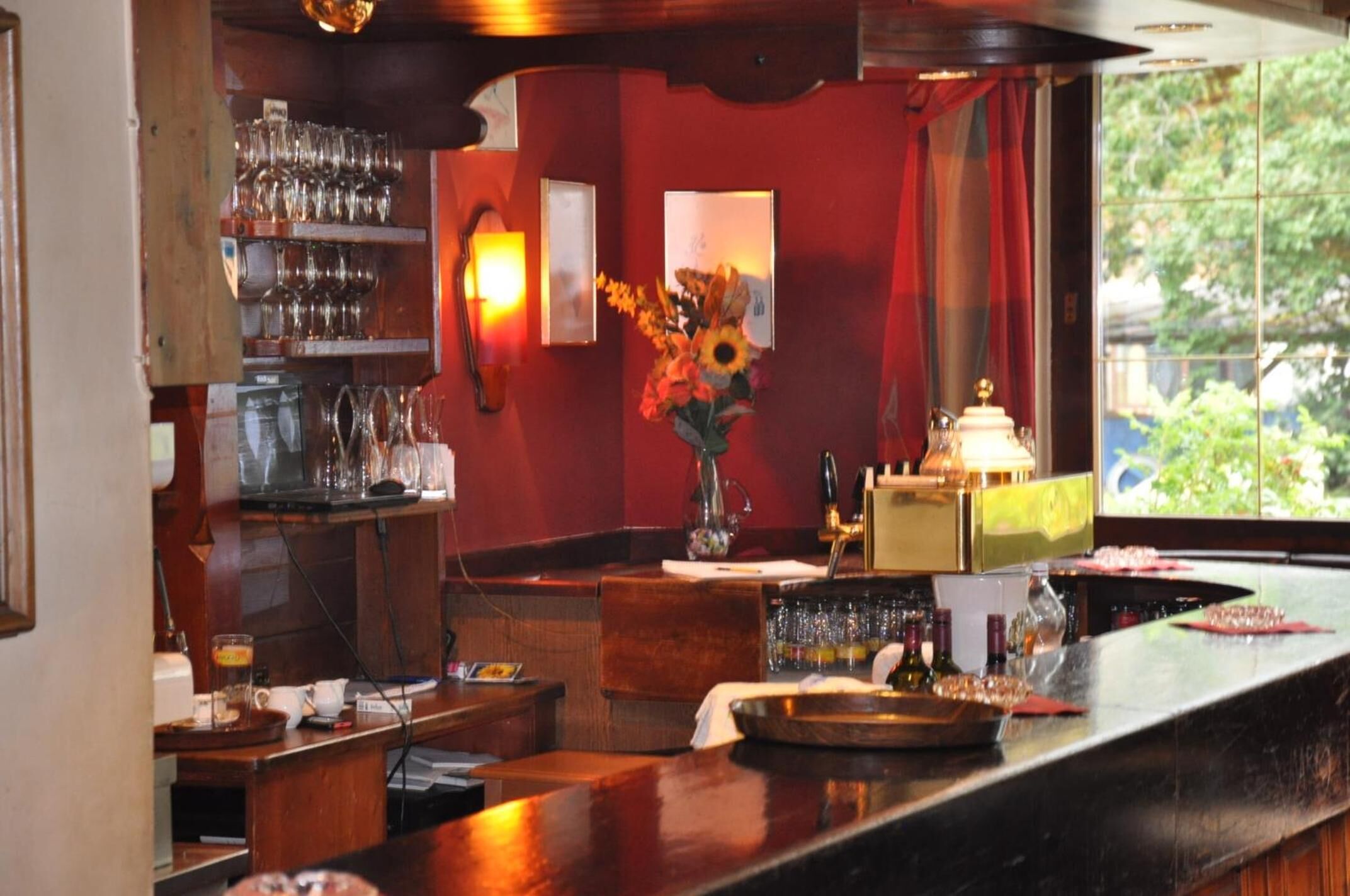 A bar area with a wooden counter, shelves with glasses, a vase with flowers, and a window with greenery outside.