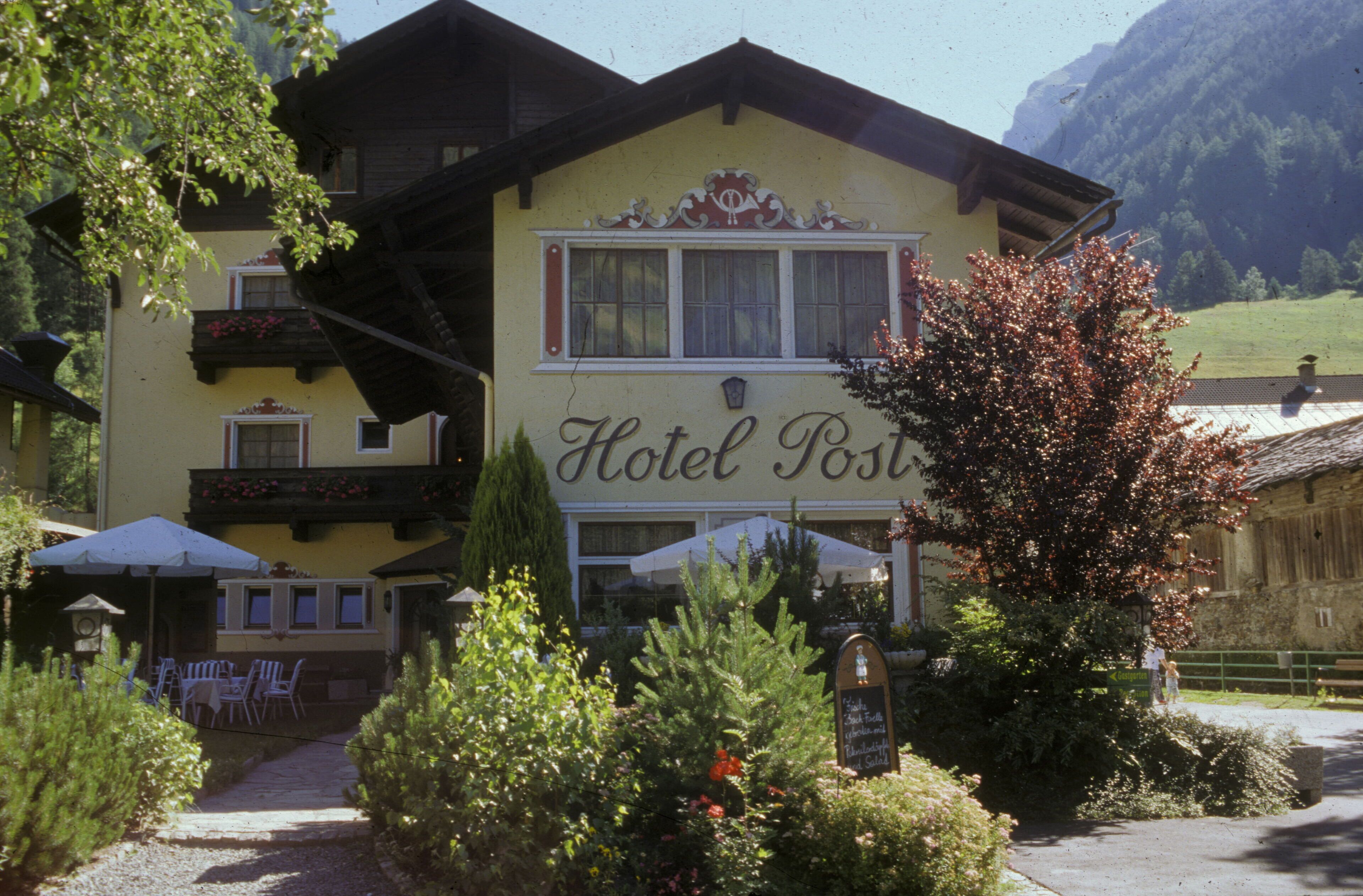 A two-story building with a sign reading "Hotel Post". There are outdoor tables and chairs, and a pathway leading to the entrance.