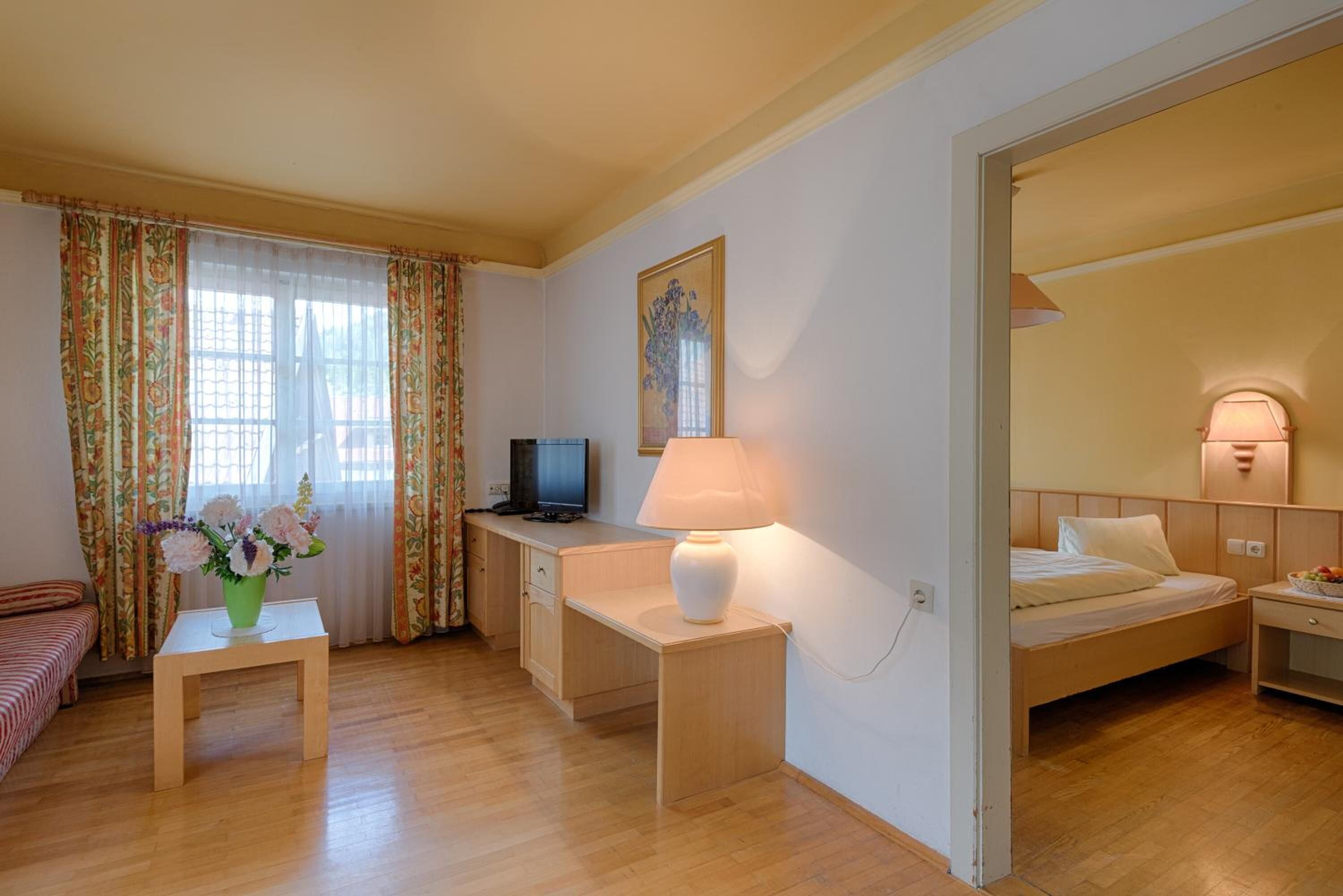 A compact hotel room with a bed, a desk with a lamp, a television, and a small table with flowers.