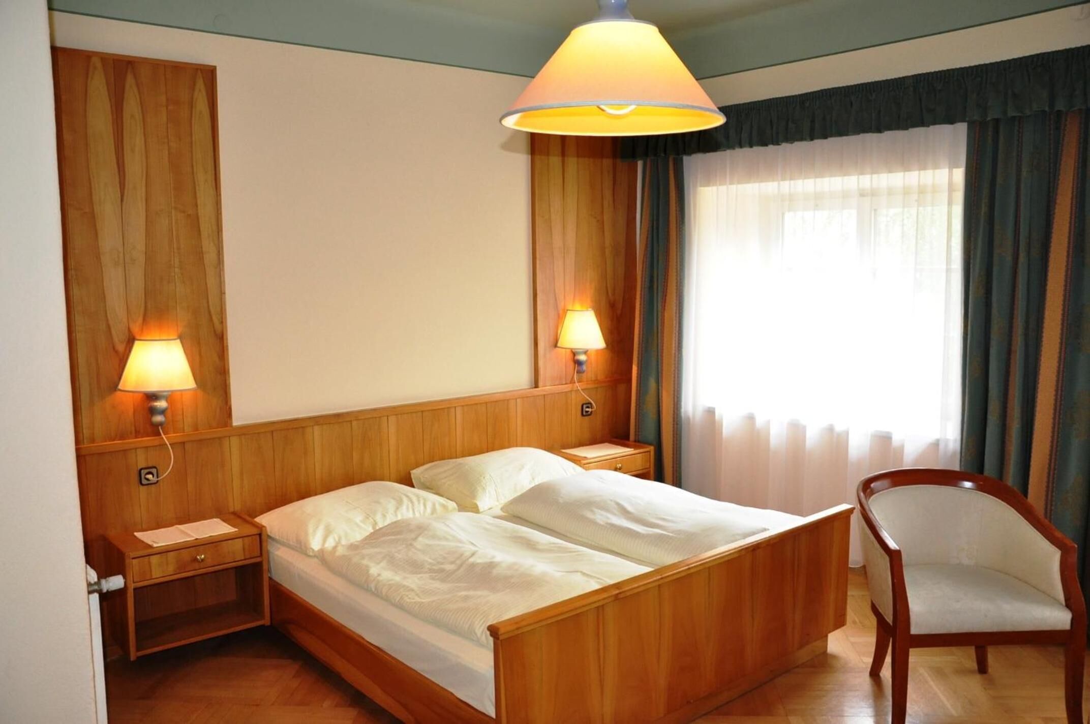 A hotel room with a wooden bed, two bedside lamps, a chair, and a large window with curtains.