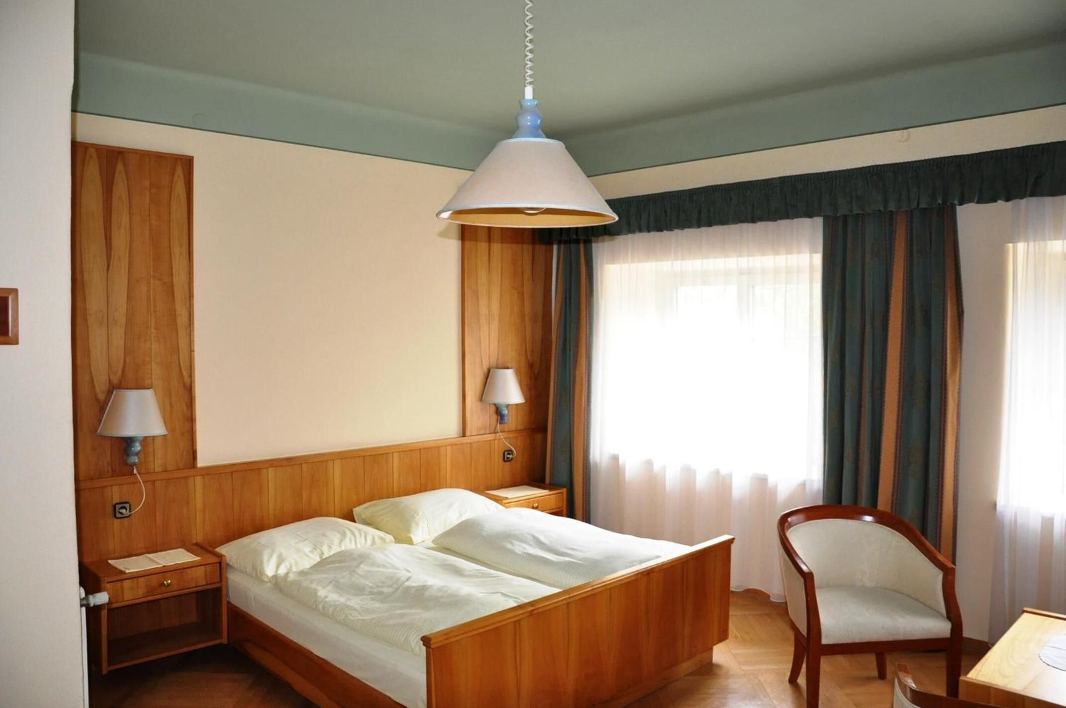 A hotel room with a wooden bed, two bedside tables with lamps, a chair, and a window with curtains.