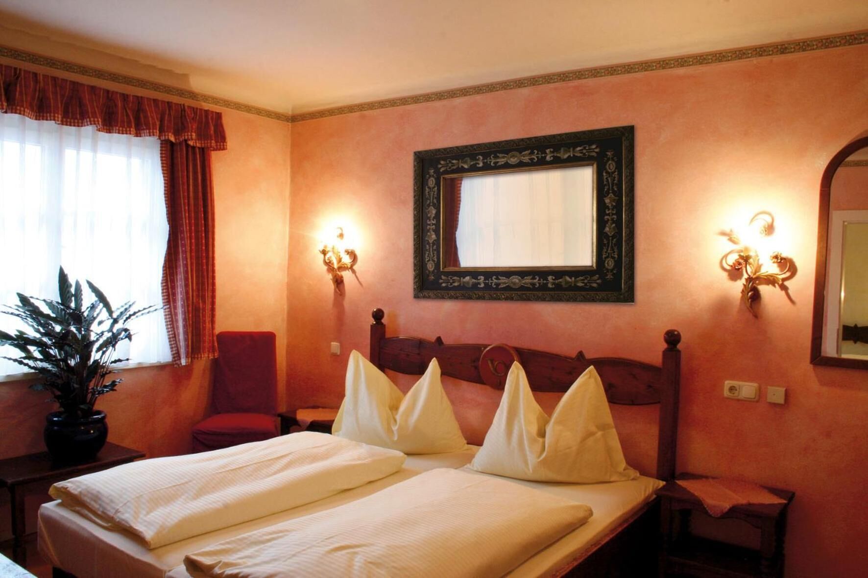 Habitación de hotel con cama doble, una silla, mesita de noche, planta en maceta y un espejo grande en la pared.