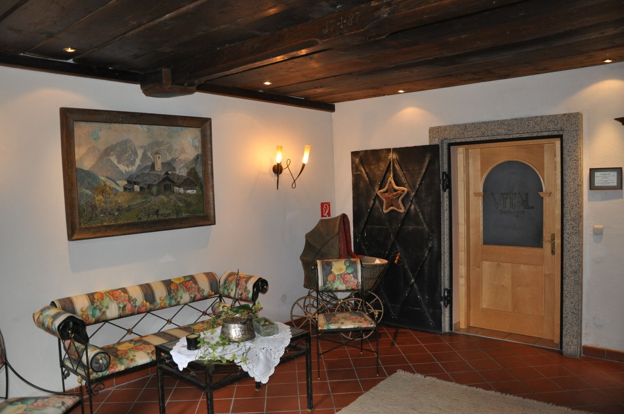 Quarto com um banco de madeira, uma pequena mesa com uma planta, um quadro na parede, uma porta com decoração em forma de estrela e um candeeiro fixado na parede.